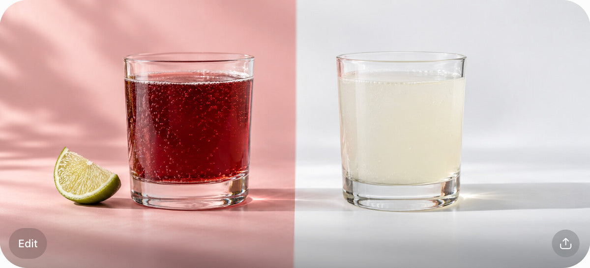 Two glasses of soda, one deep sour-cherry red with a lime wedge and one pale cloudy off-white, split on a blush and white surface